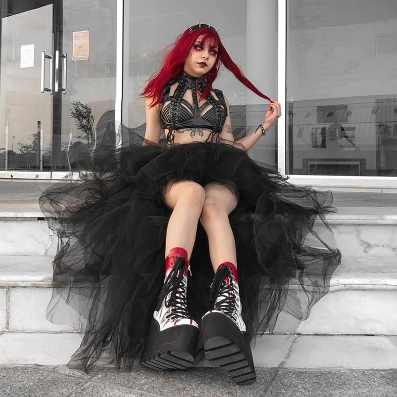 Black gothic high-low tulle skirt with flared hemline, styled with punk bustier top and chunky platform boots, seated low-angle front view on commercial building steps
