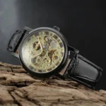 Black steampunk skeleton mechanical watch with gold hollow movement and Roman numeral dial, displayed on dark wooden surface with oblique top-down view showcasing leather strap detail