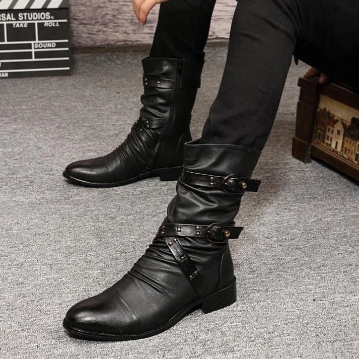 Male model wearing black steampunk boots with multiple buckle straps and ruched leather shaft in studio setting with industrial backdrop