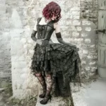 Black steampunk gothic lace cocktail dress with ruffled high-low skirt, rear-side view showing lace-up corset back and tiered lace detail on vintage stone wall background