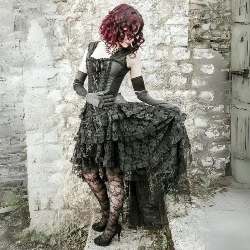 Black steampunk gothic lace cocktail dress with ruffled high-low skirt, rear-side view showing lace-up corset back and tiered lace detail on vintage stone wall background
