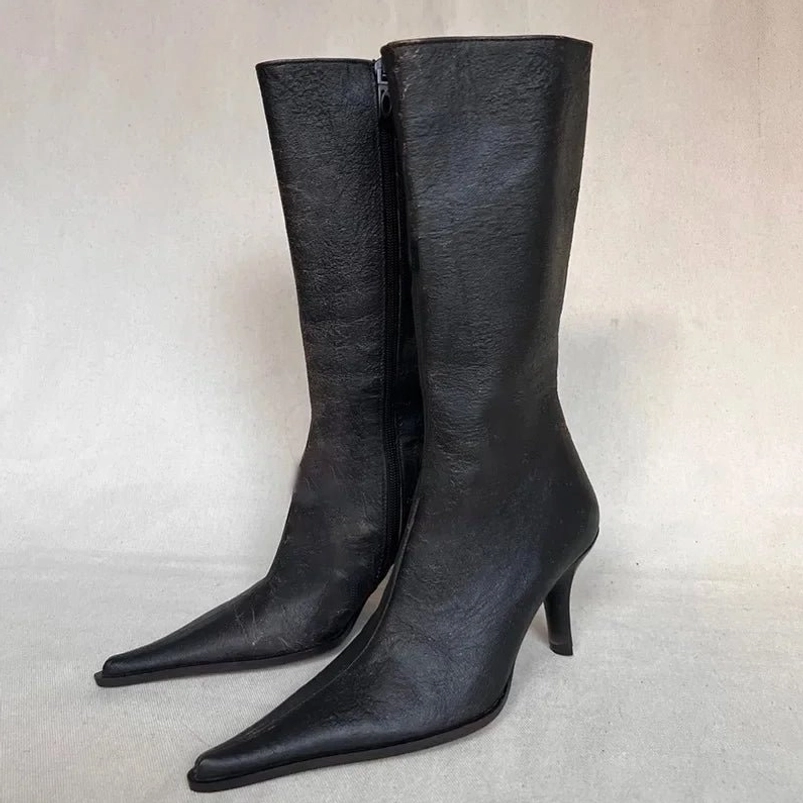 Minimalist black mid-calf stiletto boots three-quarter side view, smooth matte black leather with pointed toe and clean architectural lines