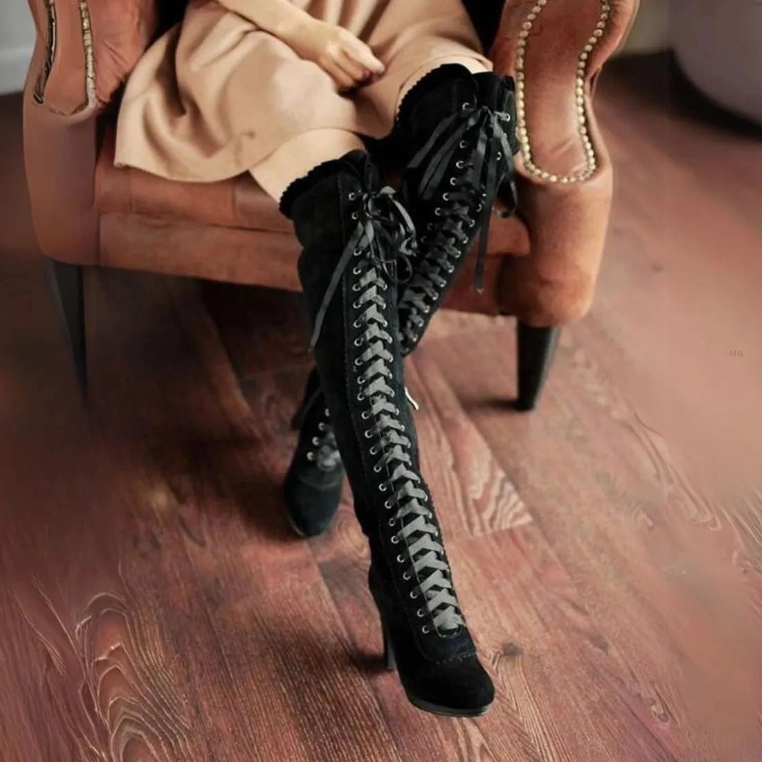 Black suede steampunk lace-up over-the-knee boots with satin ribbon lacing and ruffled collar, seated front oblique view on vintage wooden floor
