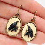 Antiqued bronze raven cameo drop earrings with black raven relief on cream base, held in hand showing close-up detail and hanging length