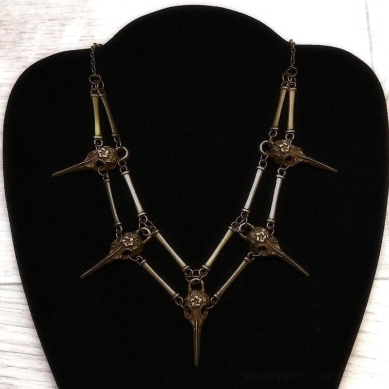 Bronze bird skull skeleton necklace with layered V-shaped design, displayed on jewelry bust against wooden surface