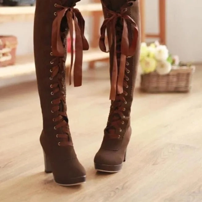 Brown suede over-the-knee steampunk lace-up boots with block high heel, front view in indoor home setting with wooden floor and flower basket