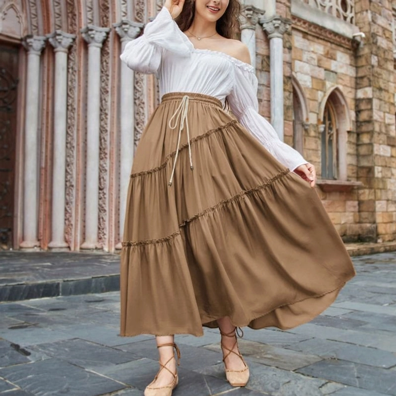 Brown tiered maxi skirt with ruffled layers worn by model in Victorian stone church courtyard, front three-quarter view showing elastic drawstring waist and cascading ruffled silhouette