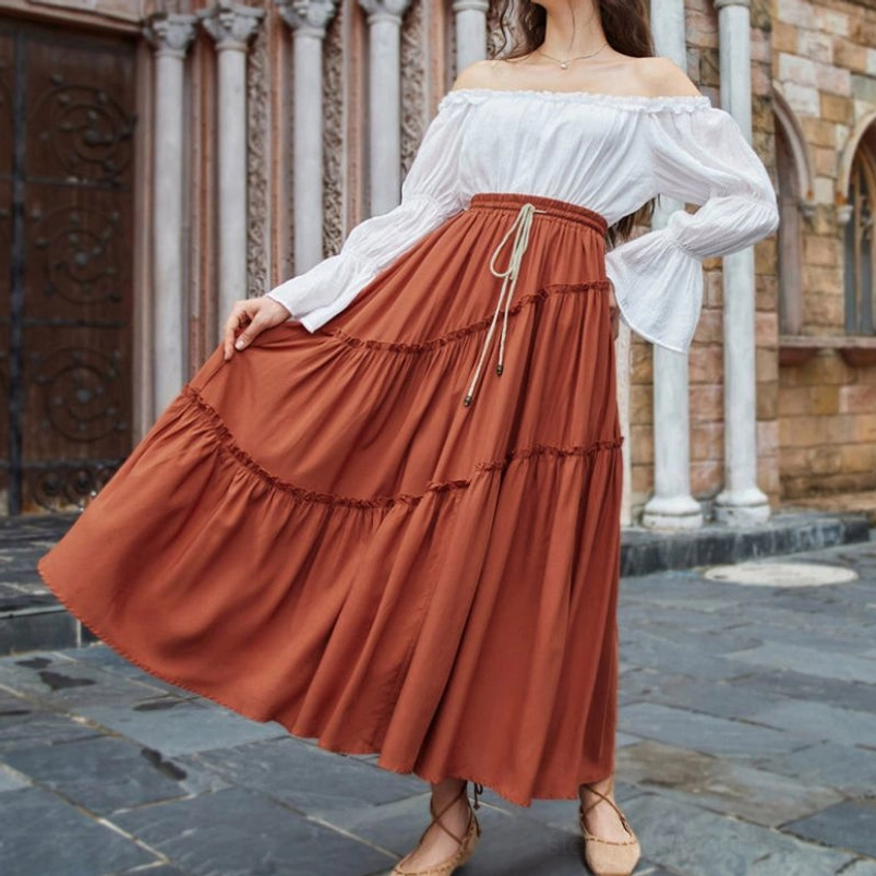 Burnt orange tiered maxi skirt worn by model in stone courtyard, front view showing drawstring waist and layered ruffled design in warm rust tone