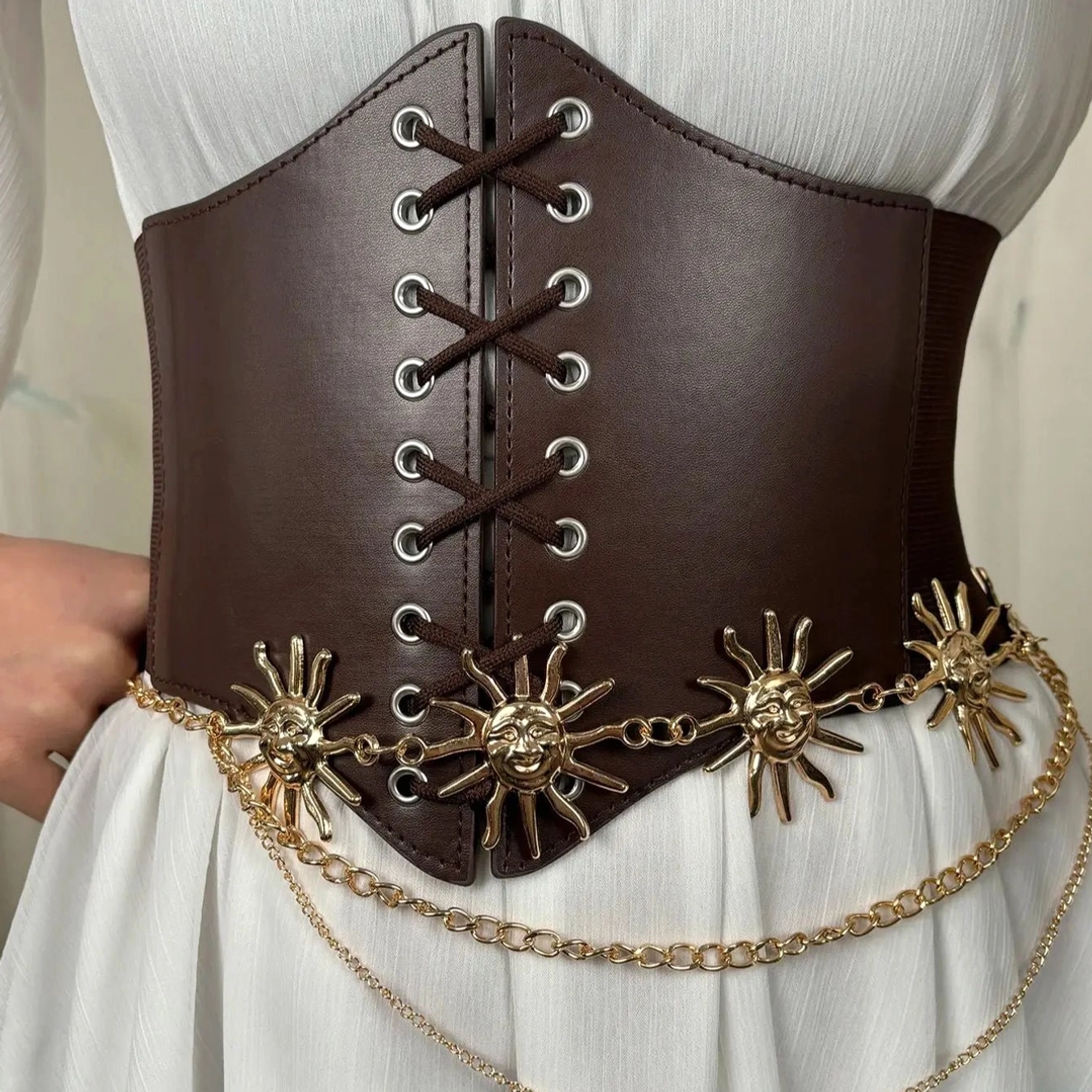 Detailed close-up of brown leather underbust corset belt showcasing precision metal eyelets, multiple intricately crafted sun-shaped metal charms, and extended layered gold chain embellishments