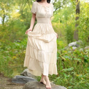 Beige steampunk maxi dress with smocked waist, tiered skirt, and short sleeves worn outdoors by model in woodland waterside setting