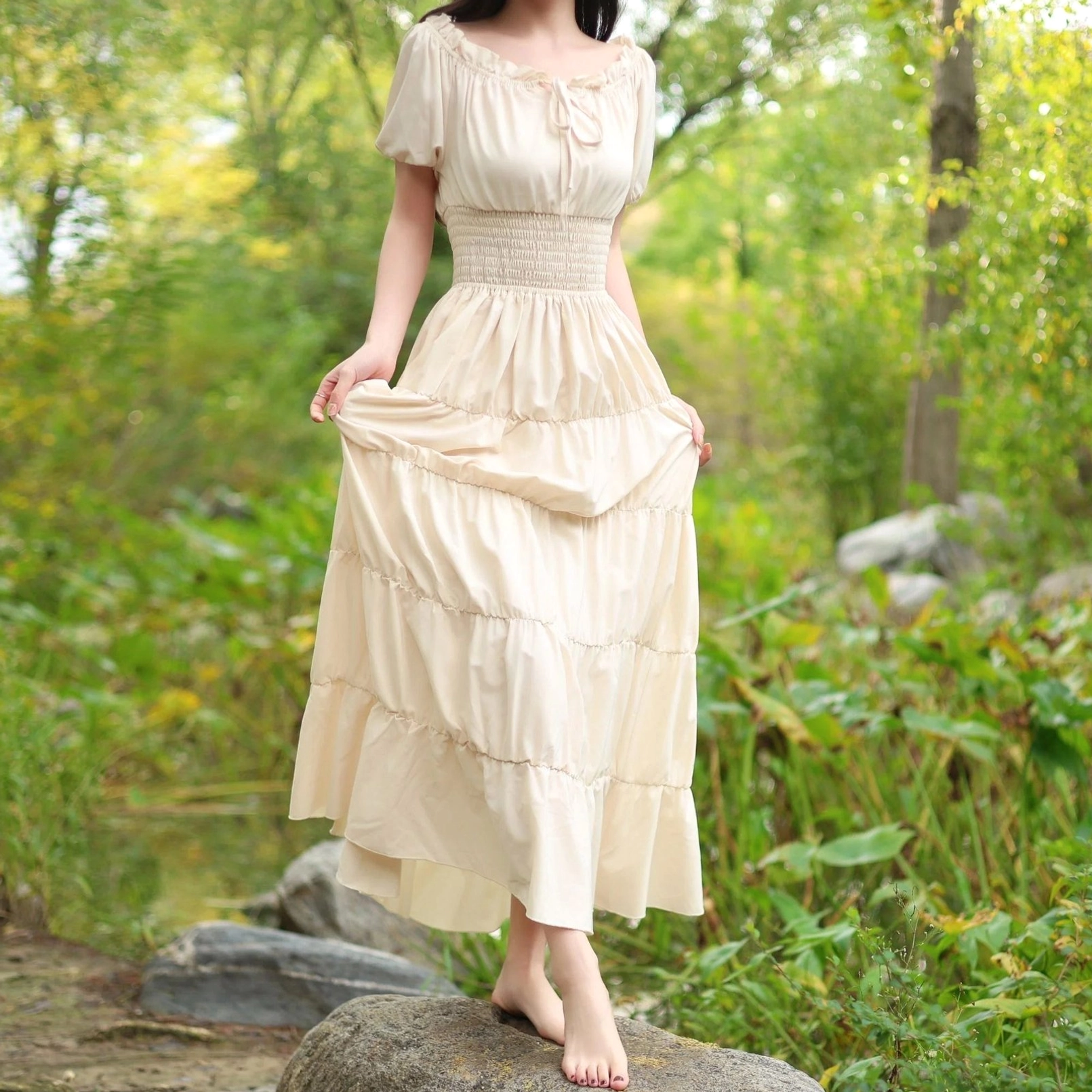 Beige steampunk maxi dress with smocked waist, tiered skirt, and short sleeves worn outdoors by model in woodland waterside setting