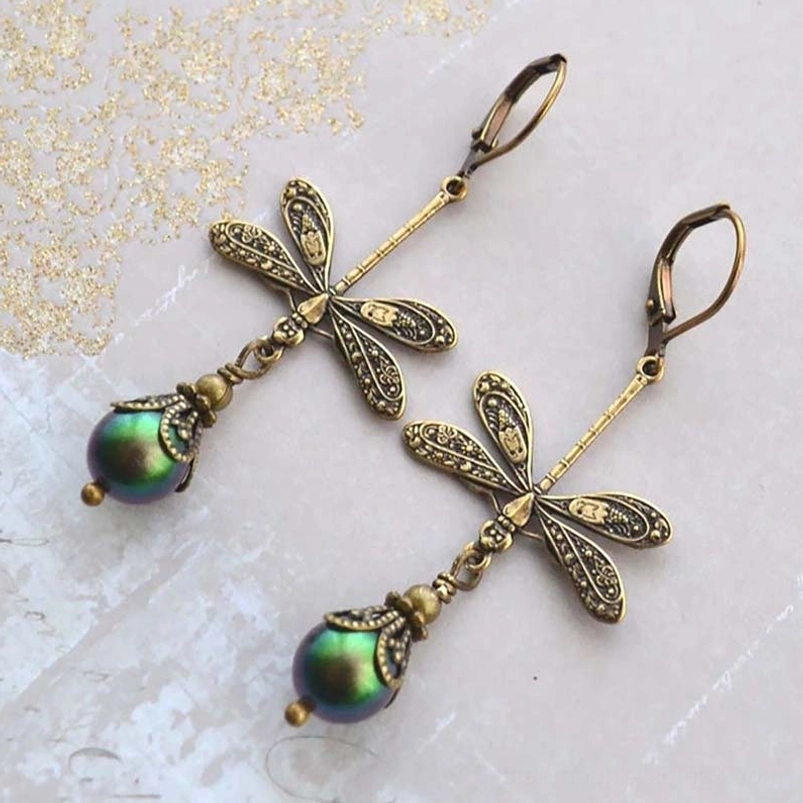 Steampunk dragonfly dangle earrings with hollow-carved antiqued bronze design and iridescent green pearl pendants displayed on vintage paper background with gold glitter patterns, oblique angle shot