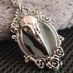 Gothic bird skull pendant necklace with vintage silver tone metal, black enamel detail, and ornate rose scroll filigree frame on fabric surface - steampunk Victorian jewelry