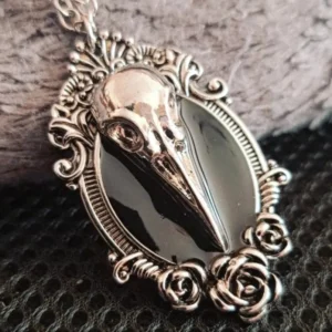 Gothic bird skull pendant necklace with vintage silver tone metal, black enamel detail, and ornate rose scroll filigree frame on fabric surface - steampunk Victorian jewelry