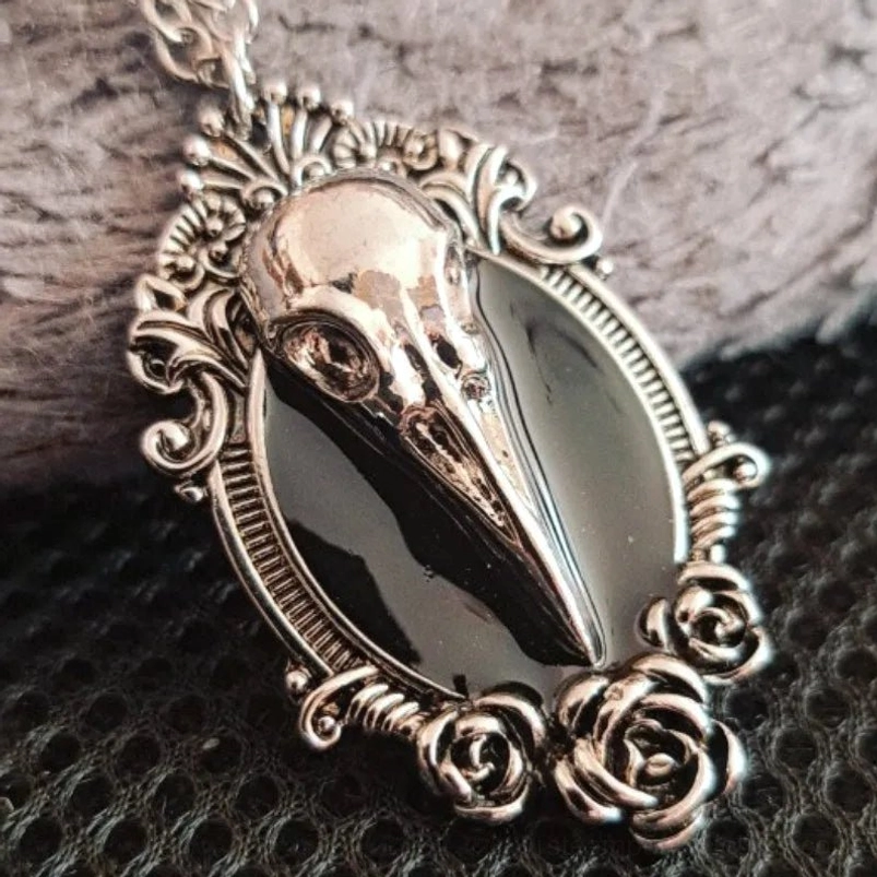 Gothic bird skull pendant necklace with vintage silver tone metal, black enamel detail, and ornate rose scroll filigree frame on fabric surface - steampunk Victorian jewelry