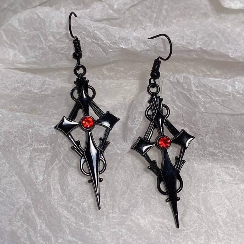 Flat lay overhead view of black gothic cross dangle earrings with ornate filigree and red rhinestone accent on fabric backdrop