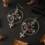 Gothic cross hoop earrings with red crystal stone and silver-tone fleur-de-lis details, displayed on dark backdrop with autumn leaves, overhead flat lay perspective
