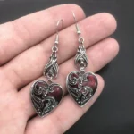 Antique silver gothic heart dangle earrings with red enamel and carved scroll details displayed on hand, close-up overhead view