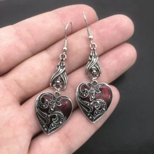 Antique silver gothic heart dangle earrings with red enamel and carved scroll details displayed on hand, close-up overhead view