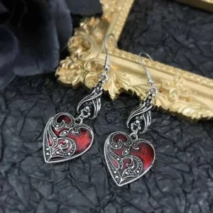 Ornate vintage gothic heart earrings with aged silver tone and deep red enamel on golden frame and dark fabric, flat lay view