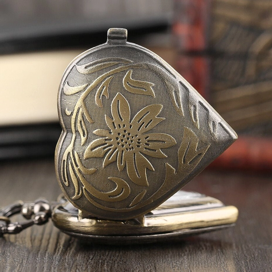 Closed heart-shaped steampunk pocket watch locket with embossed golden floral patterns on dark bronze base, three-quarter side angle view on vintage wooden desk