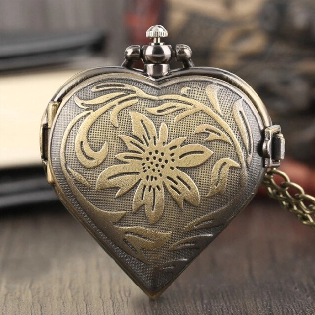 Single closed heart-shaped steampunk pocket watch featuring large embossed sunflower motif and textured metal surface, close-up front view with wooden desk and antique books background