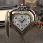 Heart-shaped steampunk pocket watch with case lid open, displaying antiqued bronze finish and engraved floral dial frame, shown on vintage wooden desk with old books