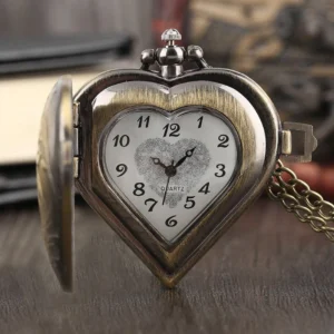 Heart-shaped steampunk pocket watch with case lid open, displaying antiqued bronze finish and engraved floral dial frame, shown on vintage wooden desk with old books