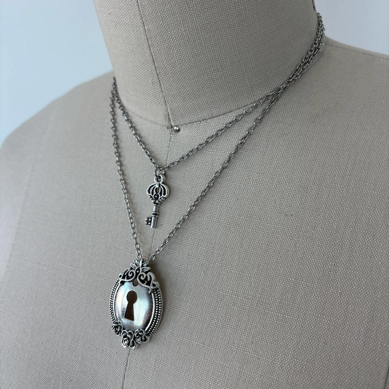 Double-strand lock and key layered necklace featuring ornate oval lock pendant with keyhole detail and silver-toned curb chain on mannequin neck