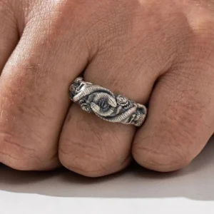 Men's gothic steampunk ring worn on middle finger, front-facing view displaying intricate double snake heads, evil eyes, and skull details with aged oxidized patina on pale studio background