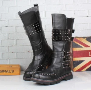 Men's black studded steampunk mid-calf combat boots with hand-positioned metal rivets and brass-tone buckle straps, genuine leather upper with patent leather accents, studio photography on white brick backdrop