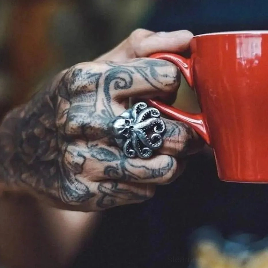 Octopus skull steampunk ring worn on tattooed male hand holding red ceramic mug, showing dark vintage metal patina in lifestyle setting