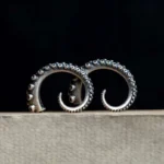 Octopus tentacle steampunk stud earrings with antiqued silver finish and detailed suction cup texture, displayed on dark backdrop with light fabric