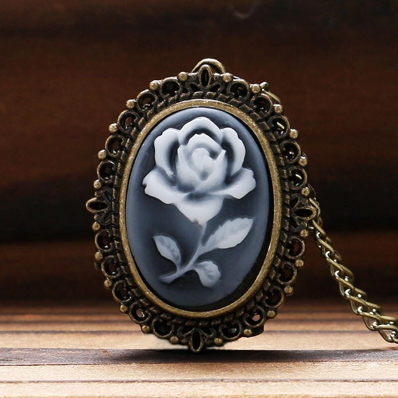 Close-up view of oval rose cameo pendant with filigree bronze frame worn on wooden surface, displaying gray-white rose design and ornate metalwork detail