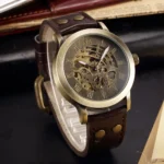 Bronze skeleton steampunk watch with brown leather strap, three-quarter view showing exposed mechanical movement and Roman numeral dial