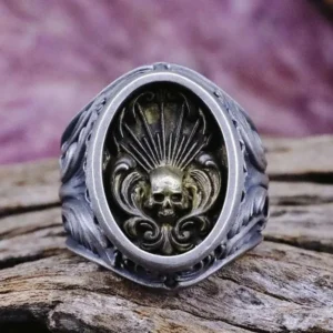 Close-up frontal view of steampunk skull ring featuring matte silver metal with detailed embossed skull motif, sunburst pattern, and ornate oval bezel frame on wooden surface