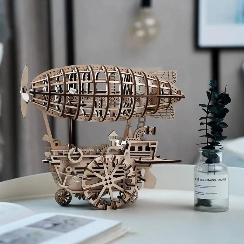 Low side angle view of steampunk wooden airship puzzle model showing detailed wood grain and mechanical structure with eucalyptus plant and open book decor