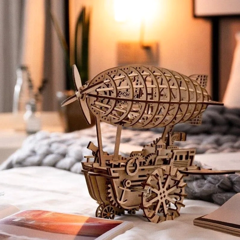 Completed steampunk airship model showing three-quarter side perspective with detailed mechanical gears and natural wood finish on warm-lit bed display