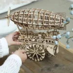 Steampunk wooden airship 3D puzzle model with laser-cut gears and mechanical framework displayed on wooden table with decorative botanicals