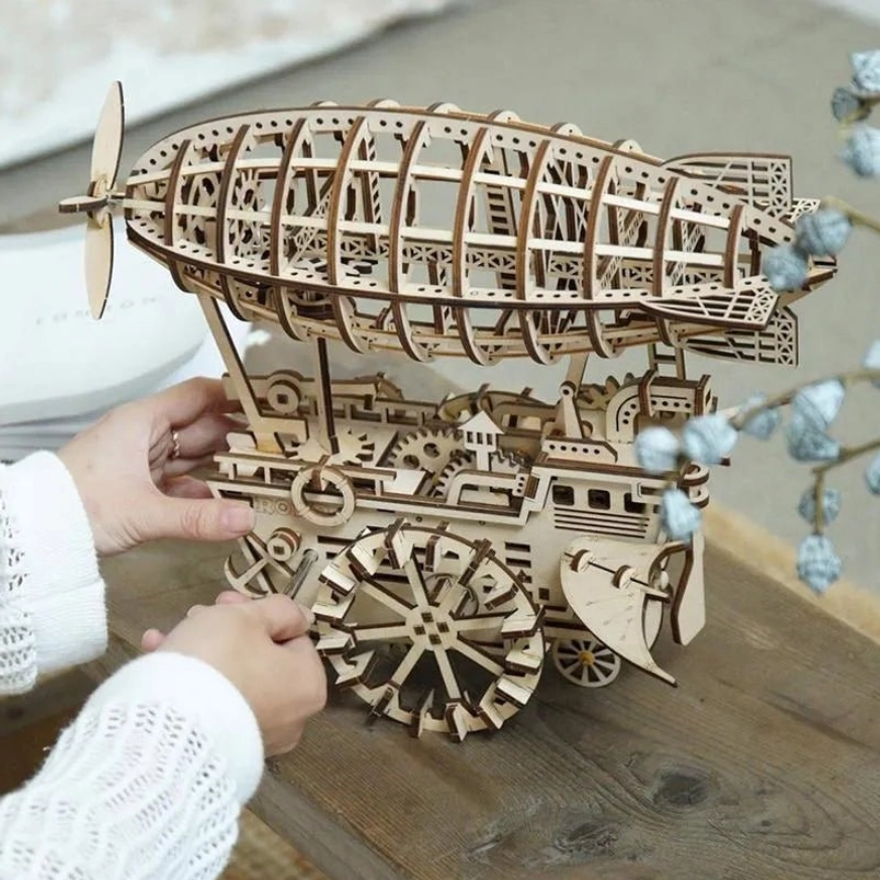 Steampunk wooden airship 3D puzzle model with laser-cut gears and mechanical framework displayed on wooden table with decorative botanicals