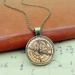 Steampunk astrolabe pendant necklace with bronze tone metal dial and glass cover, displayed on vintage leather and sheet music for authentic industrial aesthetic