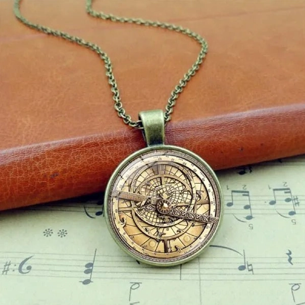 Steampunk astrolabe pendant necklace with bronze tone metal dial and glass cover, displayed on vintage leather and sheet music for authentic industrial aesthetic