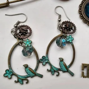 Steampunk bird loop earrings with verdigris bronze frame, intricate gears, blue crystal beads, and delicate clover accents displayed in flat lay with spare jewelry components