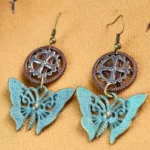 Steampunk butterfly gear drop earrings featuring patina blue hollow-carved butterfly charms with vintage bronze gear pendants on brown fabric background, flat lay overhead view showing intricate mechanical design