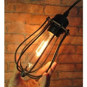Steampunk industrial Edison cage pendant lamp with distressed black metal frame and warm glowing vintage bulb, close-up side view against brick wall backdrop