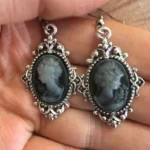Steampunk cameo drop earrings with ornate silver filigree frame and grey-white female portrait, displayed in palm with close-up detail view