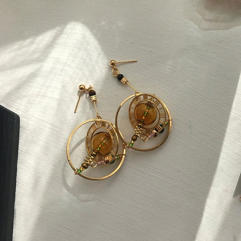 Steampunk celestial orbit earrings top-down view on white wooden table with natural light, gold hollow design with amber glass and colorful tiny beaded accents