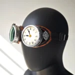 Steampunk clock goggles with aged copper trim and green tinted polycarbonate lenses, featuring Roman numeral clock decoration and compass accent, worn on black mannequin head against gray background