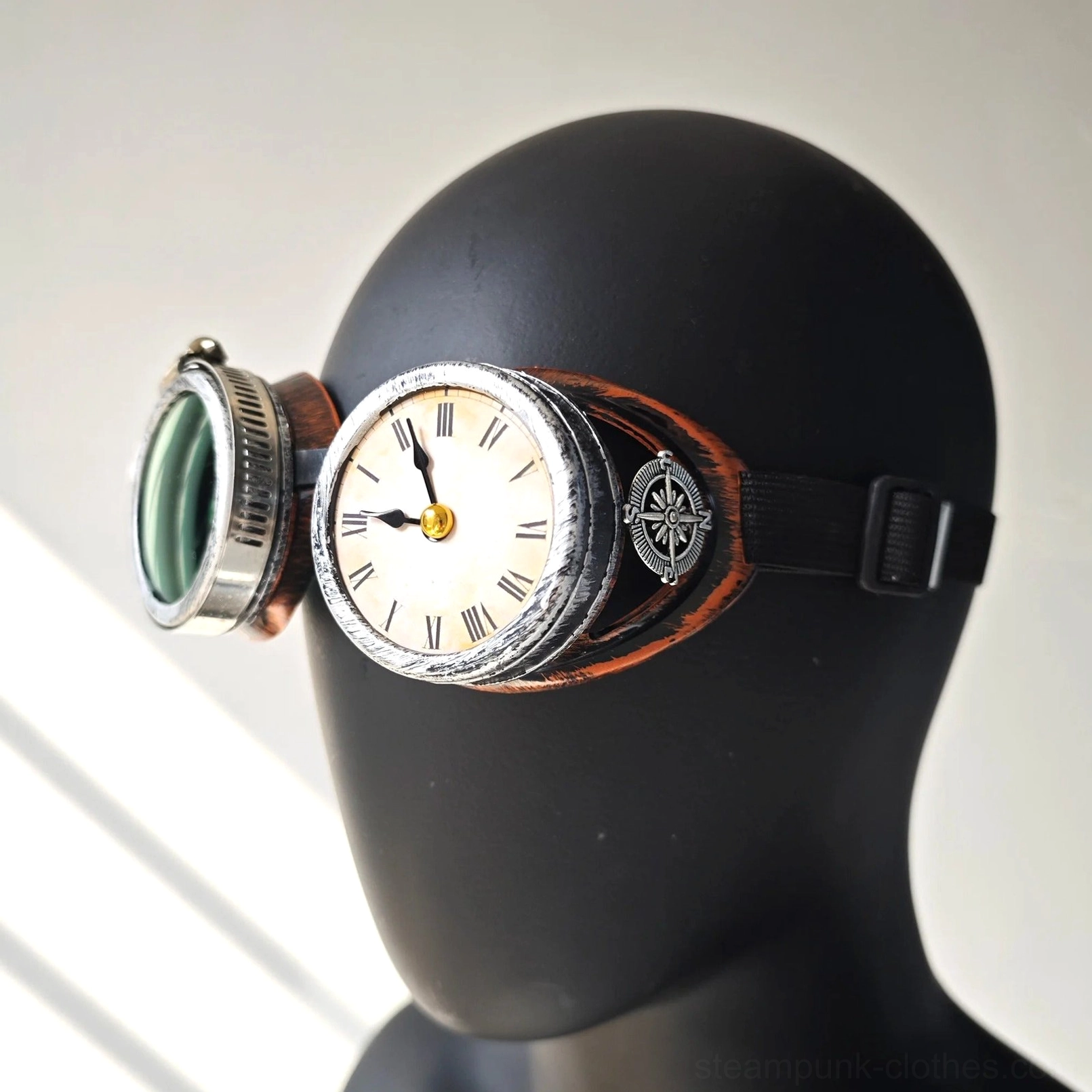 Steampunk clock goggles with aged copper trim and green tinted polycarbonate lenses, featuring Roman numeral clock decoration and compass accent, worn on black mannequin head against gray background