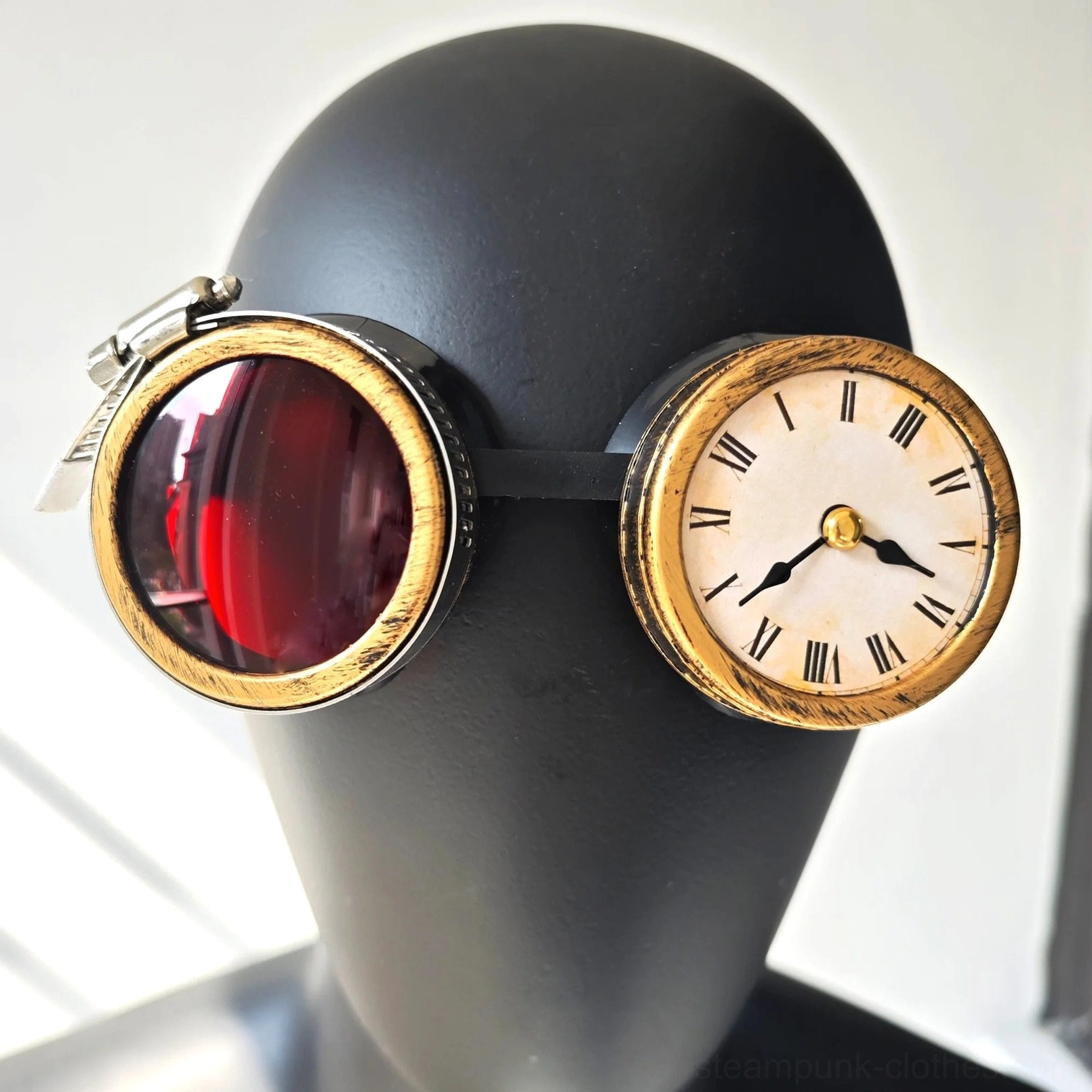 Steampunk clock goggles with gold-toned frame, red-tinted polycarbonate lenses, rotating flip-up lens cap, vintage clock decoration, and compass accents on black mannequin head